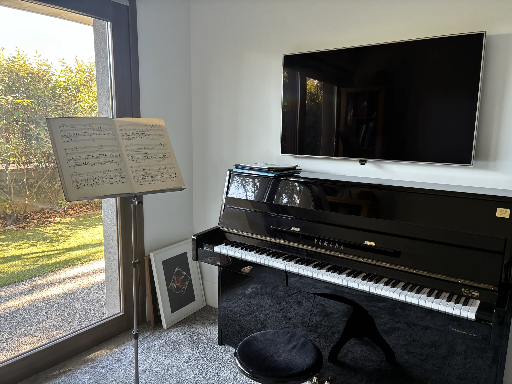 Le studio d'Élodie Tisserand : un piano droit, des pupitres et des partitions devant une grande baie vitrée ouverte sur le jardin.