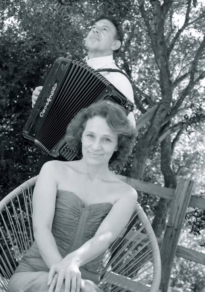 Photographie en noir et blanc : Élodie Tisserand assise en plein air, Armand Paoli debout derrière elle à l'accordéon, sous les arbres.